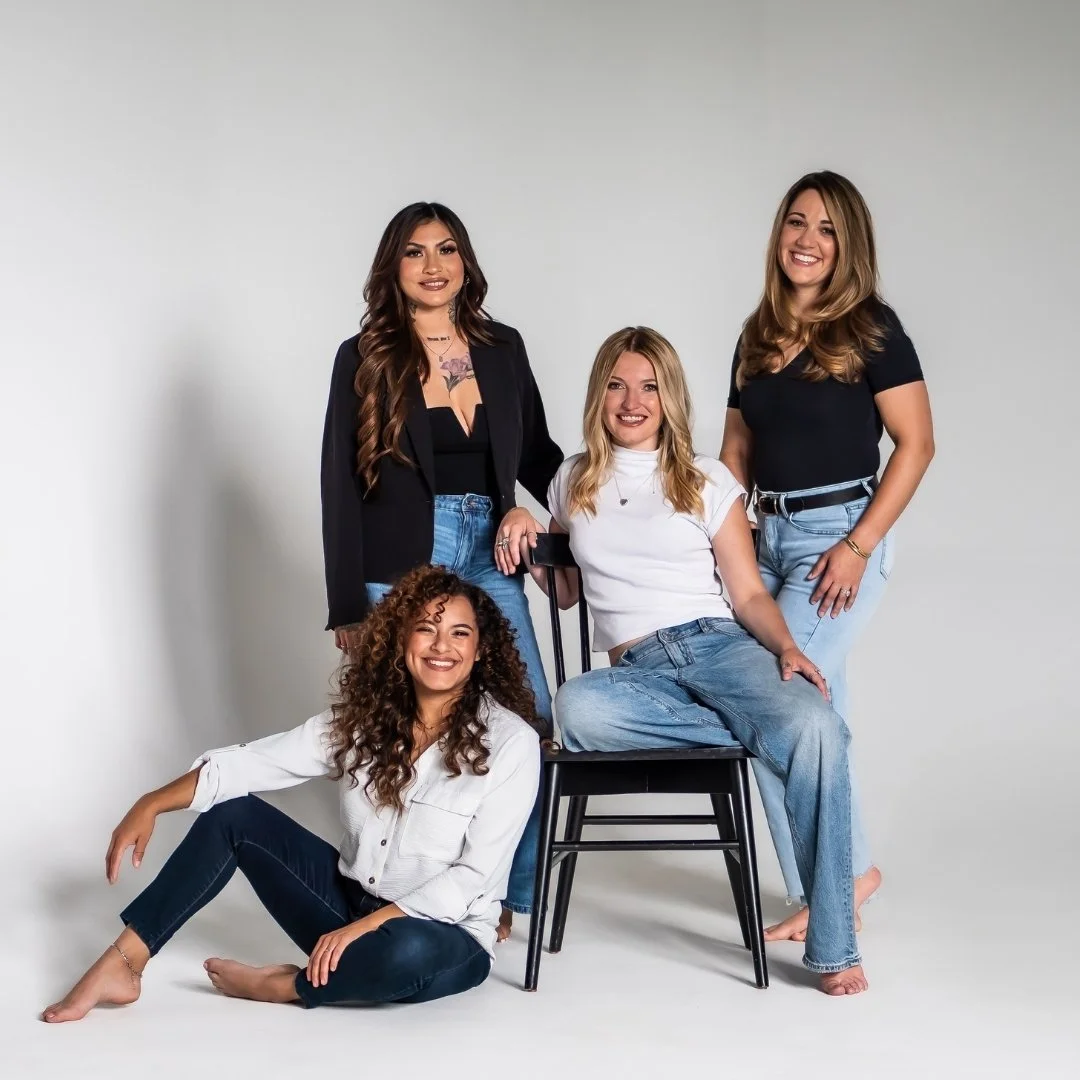 Group of five women smiling, posing together in casual clothes against a white background.
