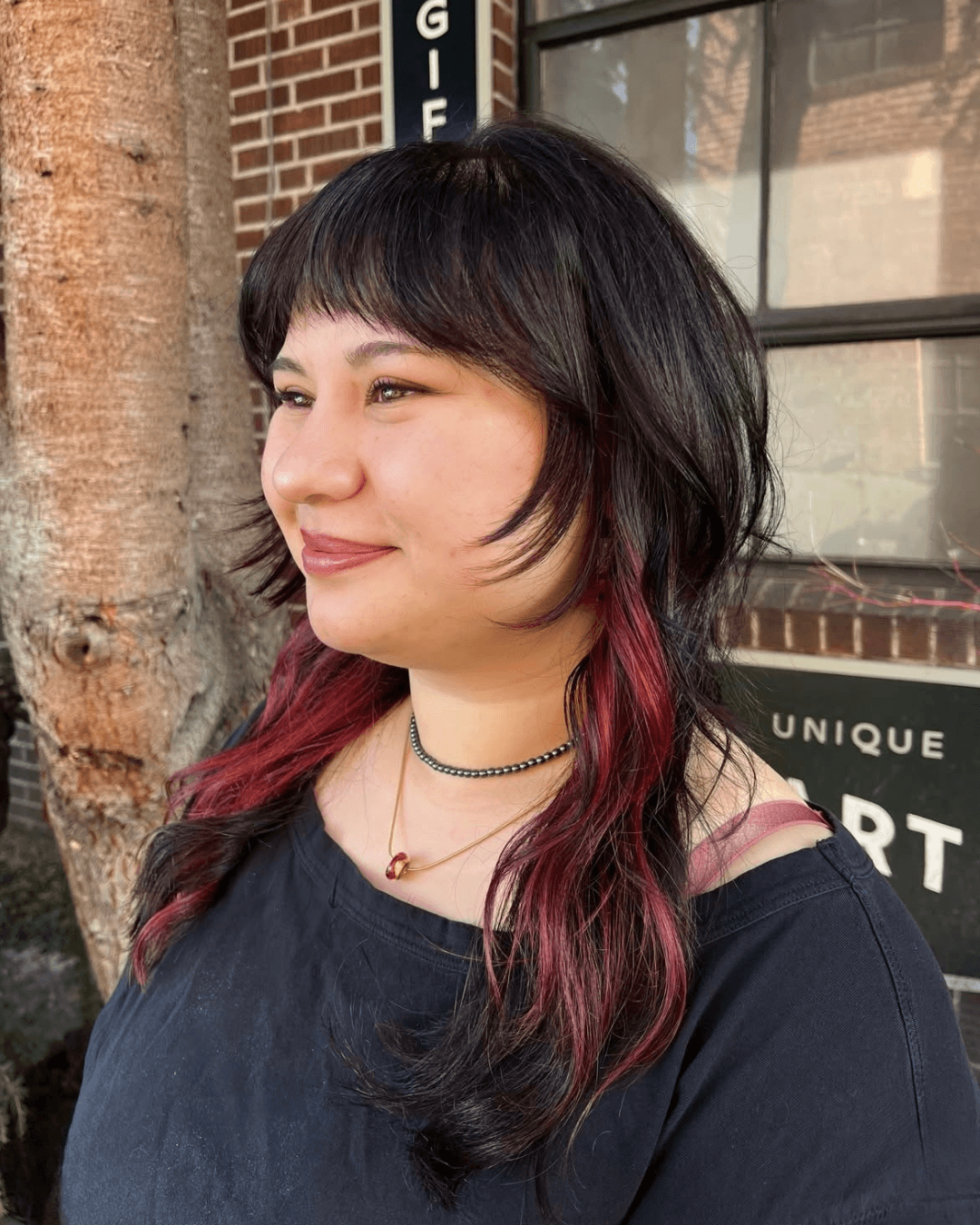 A woman with shoulder-length black hair and pink-tinted ends, wearing a black top, a black beaded choker, and a gold necklace with a red gemstone, smiling outdoors next to a tree and a brick building.