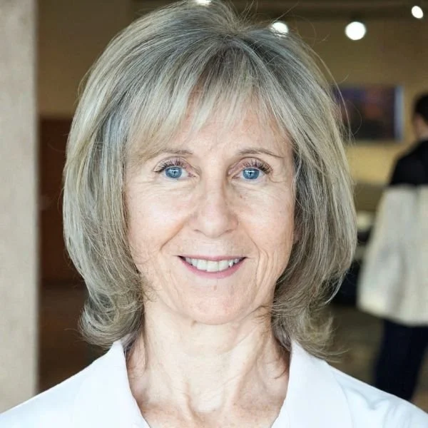 Close-up of a smiling middle-aged woman with blonde hair and blue eyes, wearing a white shirt, indoors with blurred background.