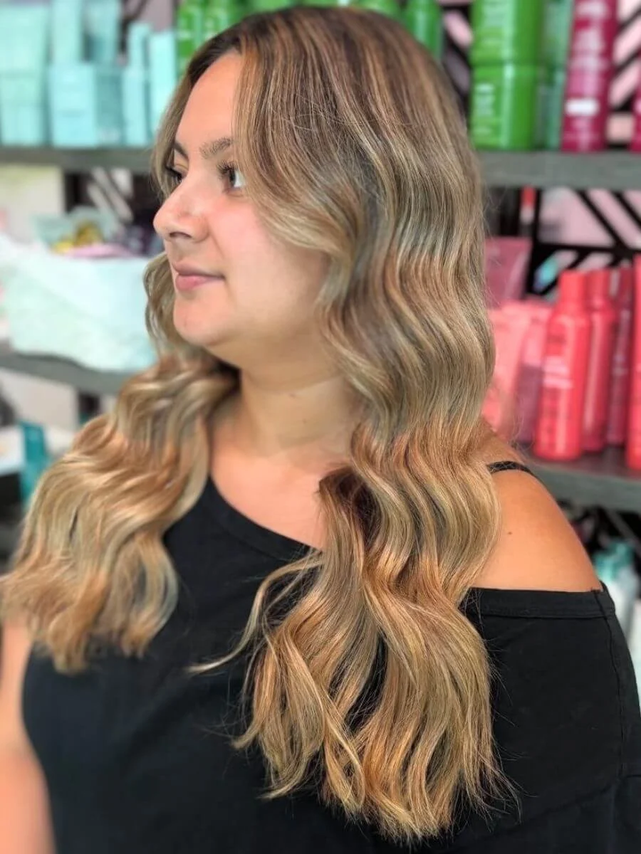 Woman with long, wavy, light brown hair in a salon or store, with shelves of colorful hair products in the background.