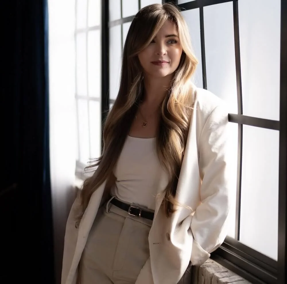 A woman with long, wavy blonde hair standing by a window with black framing, wearing a white blazer, white top, and white pants, looking slightly to the side.