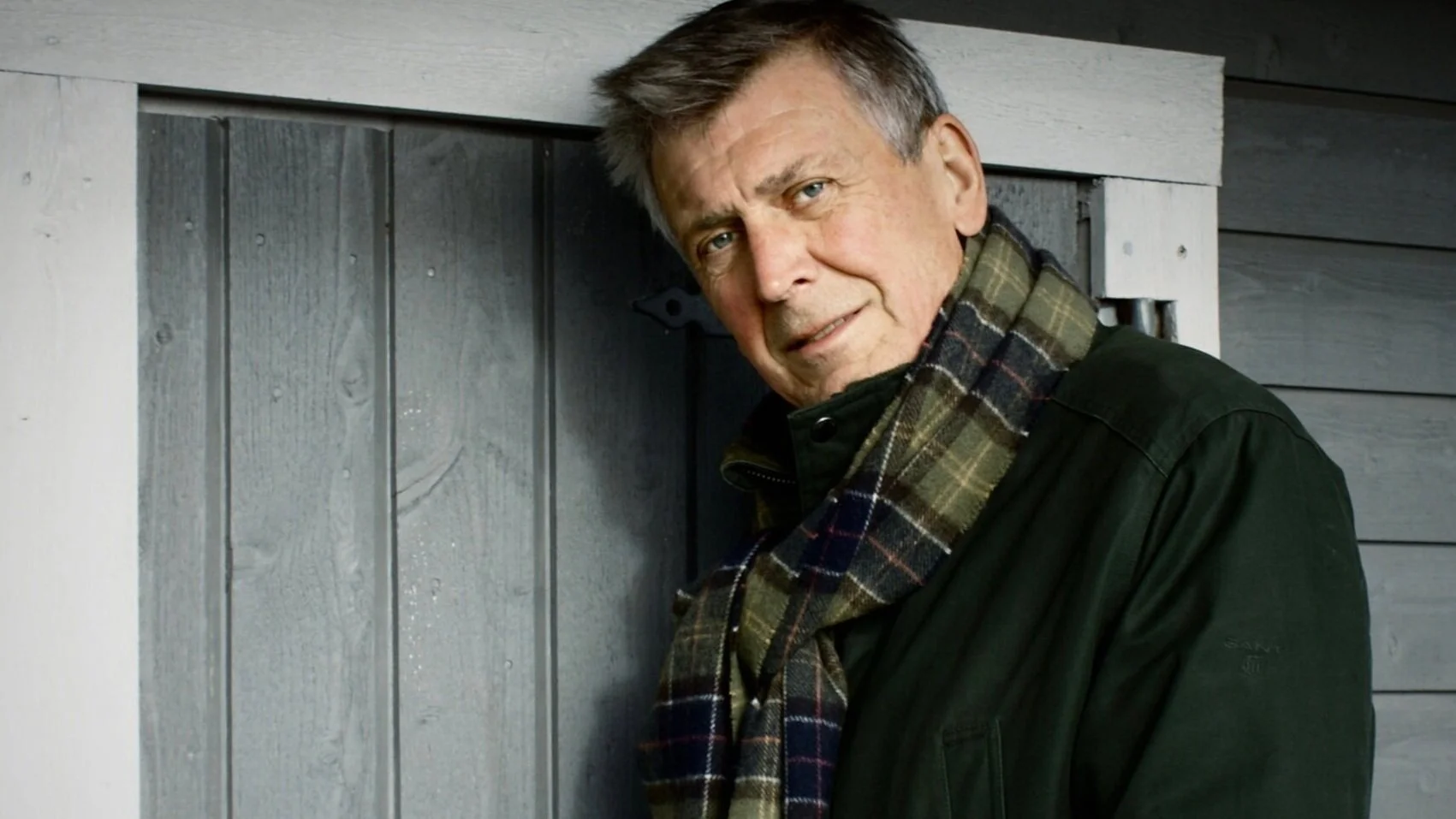 A man with gray hair, wearing a green jacket and a plaid scarf, leaning against a gray wooden wall.