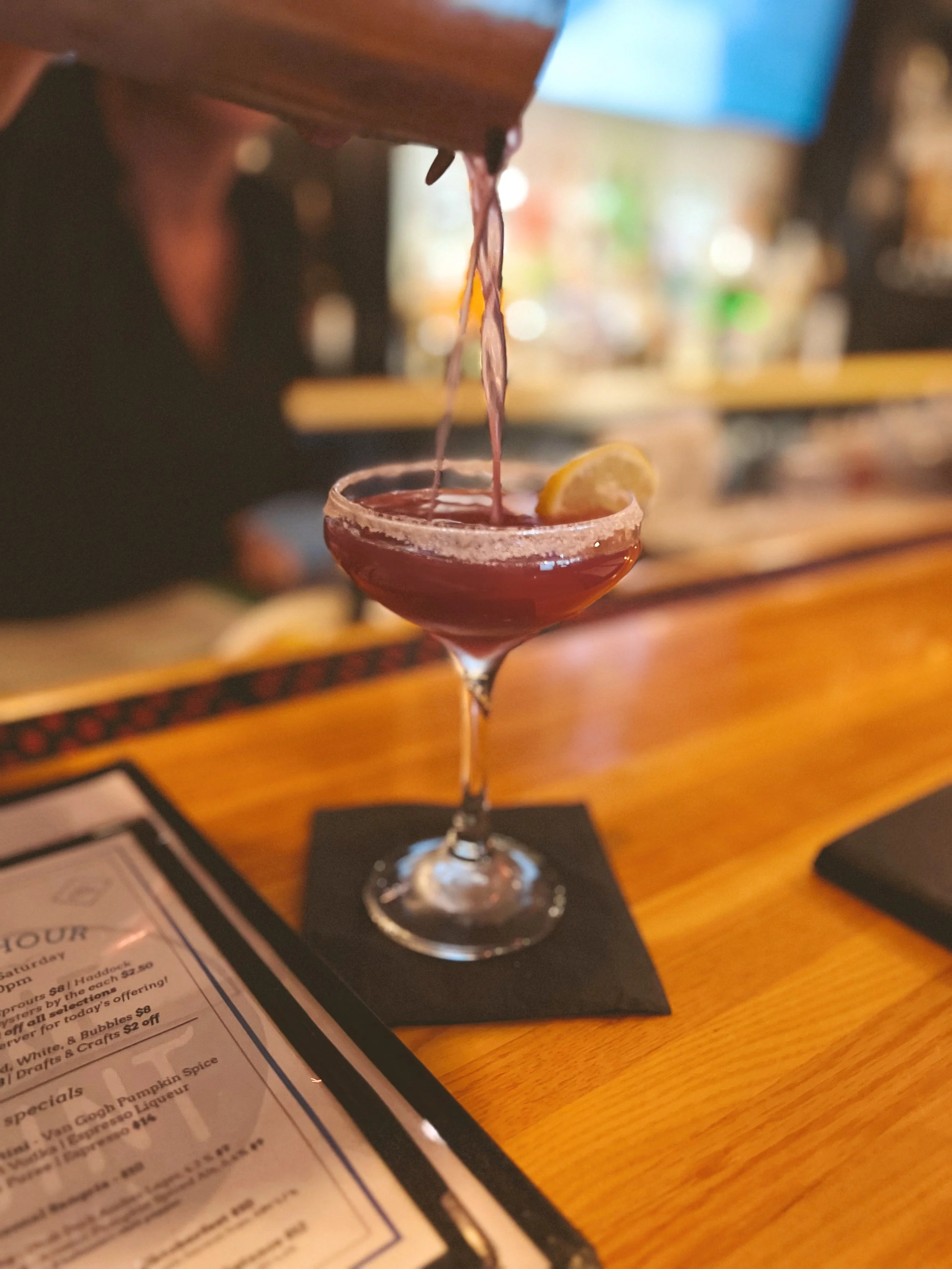 A bartender pouring a blueberry lemon drop into a martini glass with a lemon wedge garnish.
