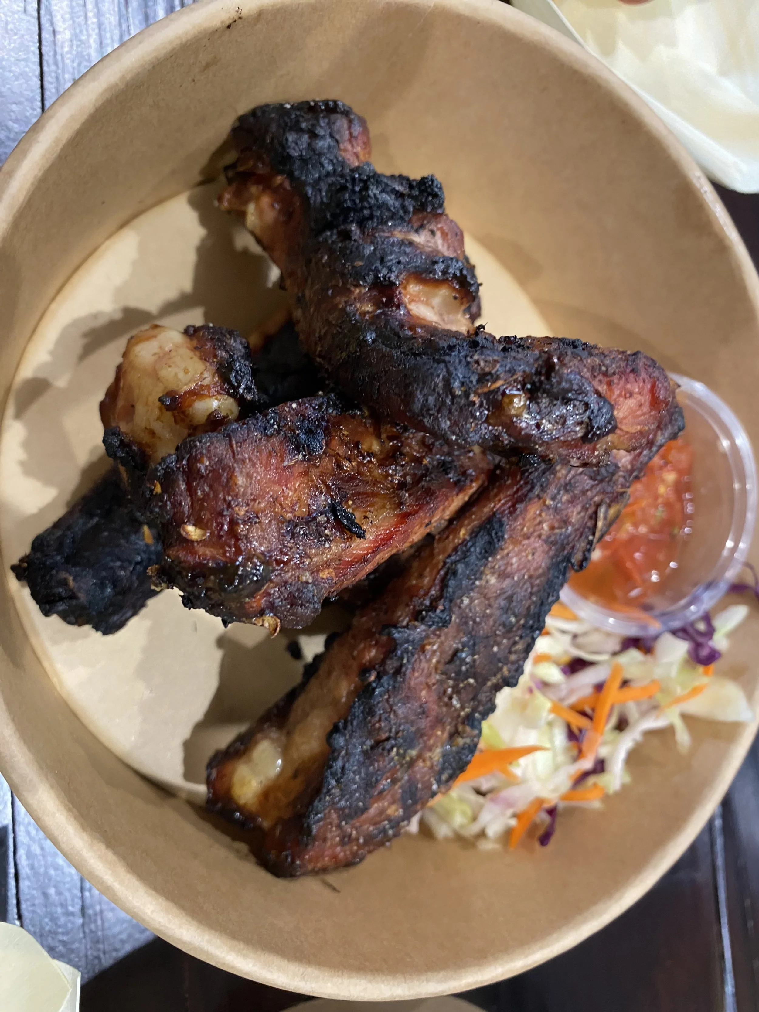 Four grilled chicken drumsticks with a charred exterior, served with a side of shredded cabbage salad and a small cup of red salsa in a brown bowl.
