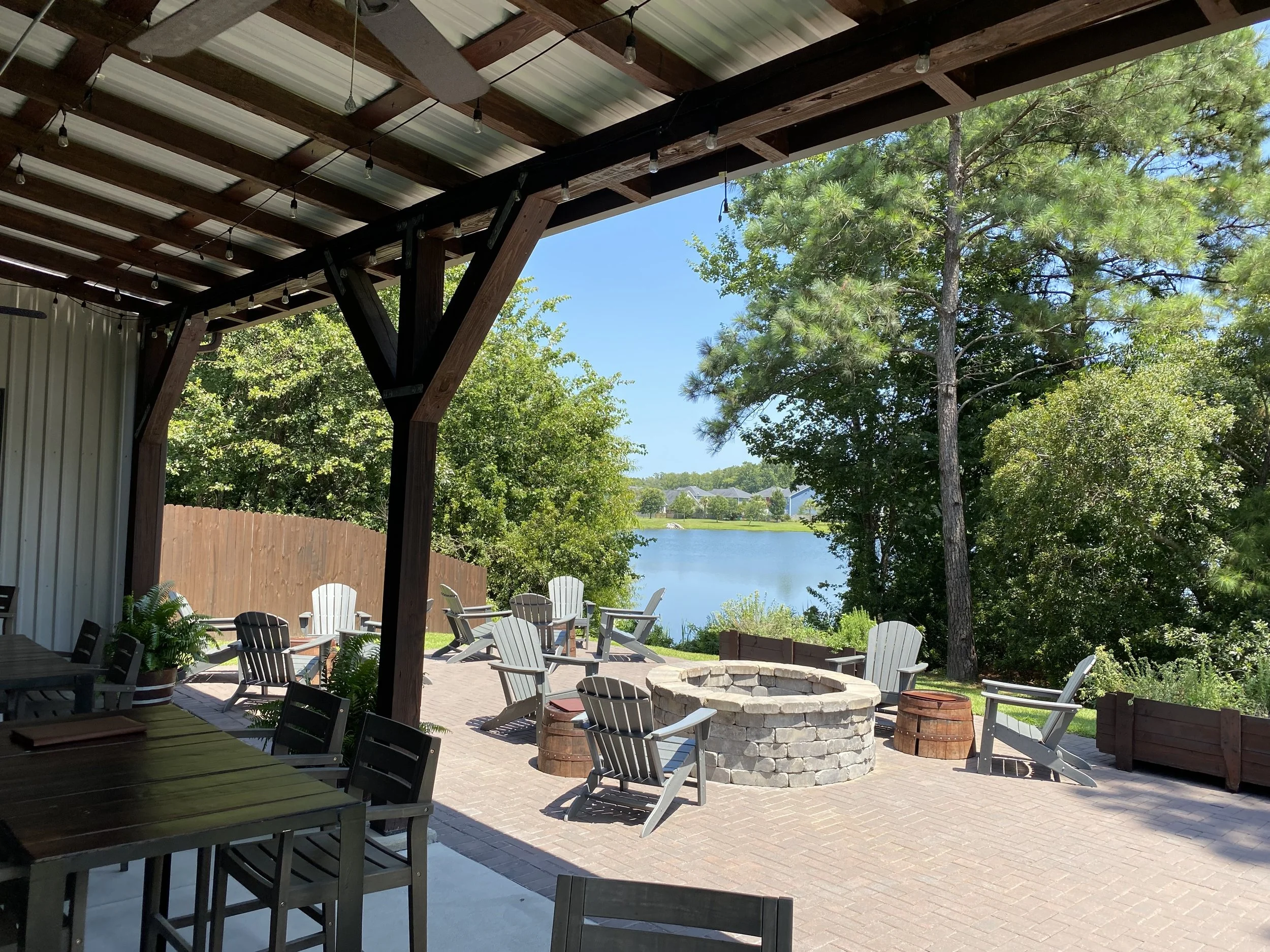 Outdoor seating area with wooden chairs and a fire pit, covered by a pergola, overlooking a scenic lake with surrounding trees.
