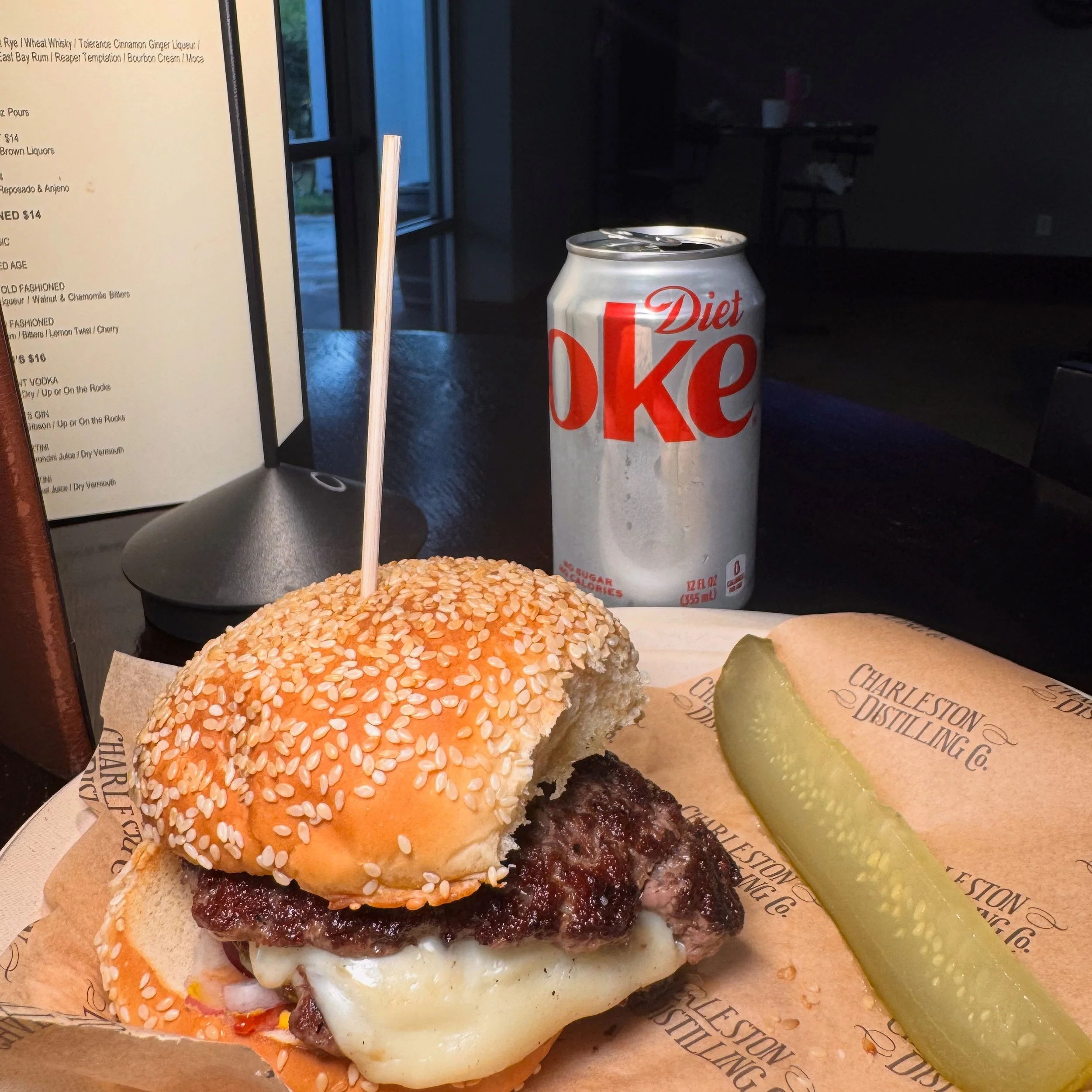 A cheeseburger with a sesame seed bun, melted cheese, and beef patty, a pickle spear on branded paper, a slice of pickle, a can of Diet Coke, and a black table and window in the background.