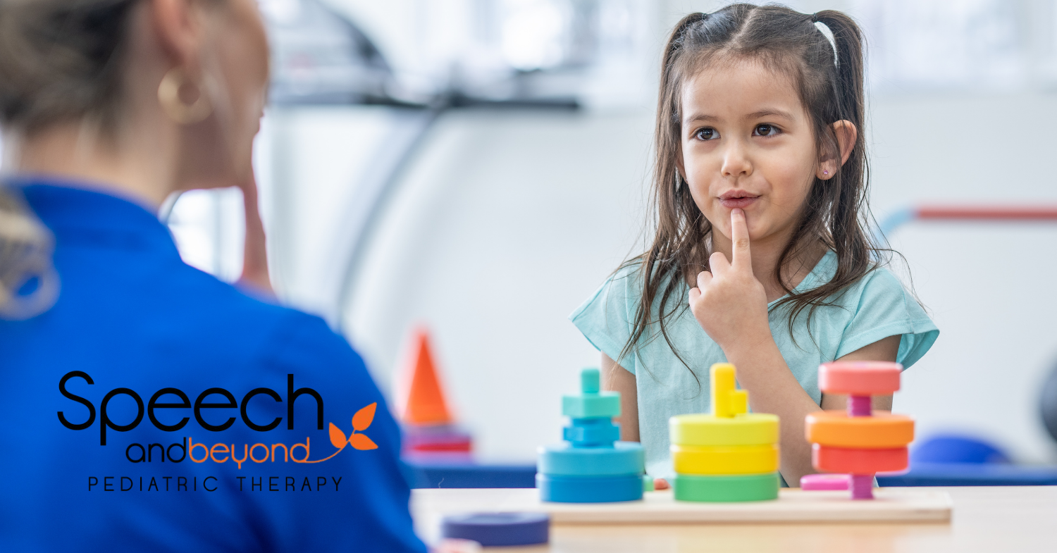 a little girl with brown hair pointing to her mouth while saying something, back of a therapist in the foreground is also pointing to her mouth