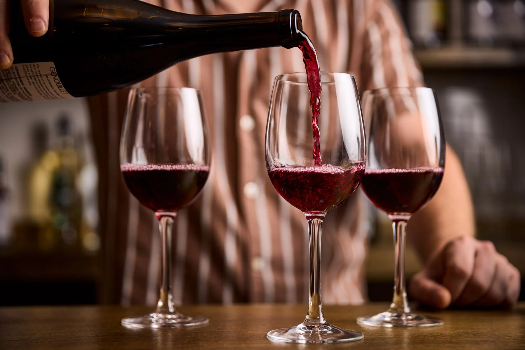 A person is pouring red wine into a wine glass with two other filled glasses on a wooden surface. The background is blurred, suggesting the bar or kitchen setting of Tria.
