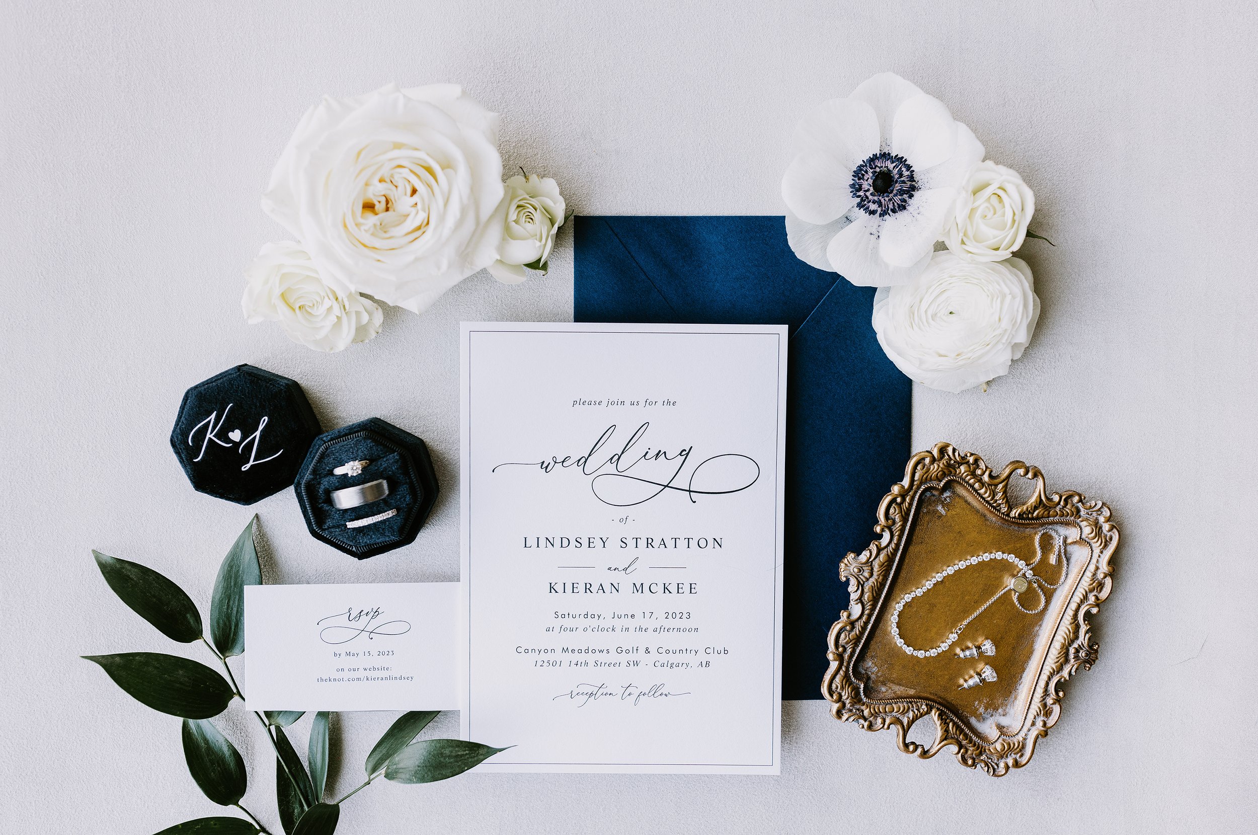 A styled wedding flat lay with an invitation, rings, jewelry and flowers