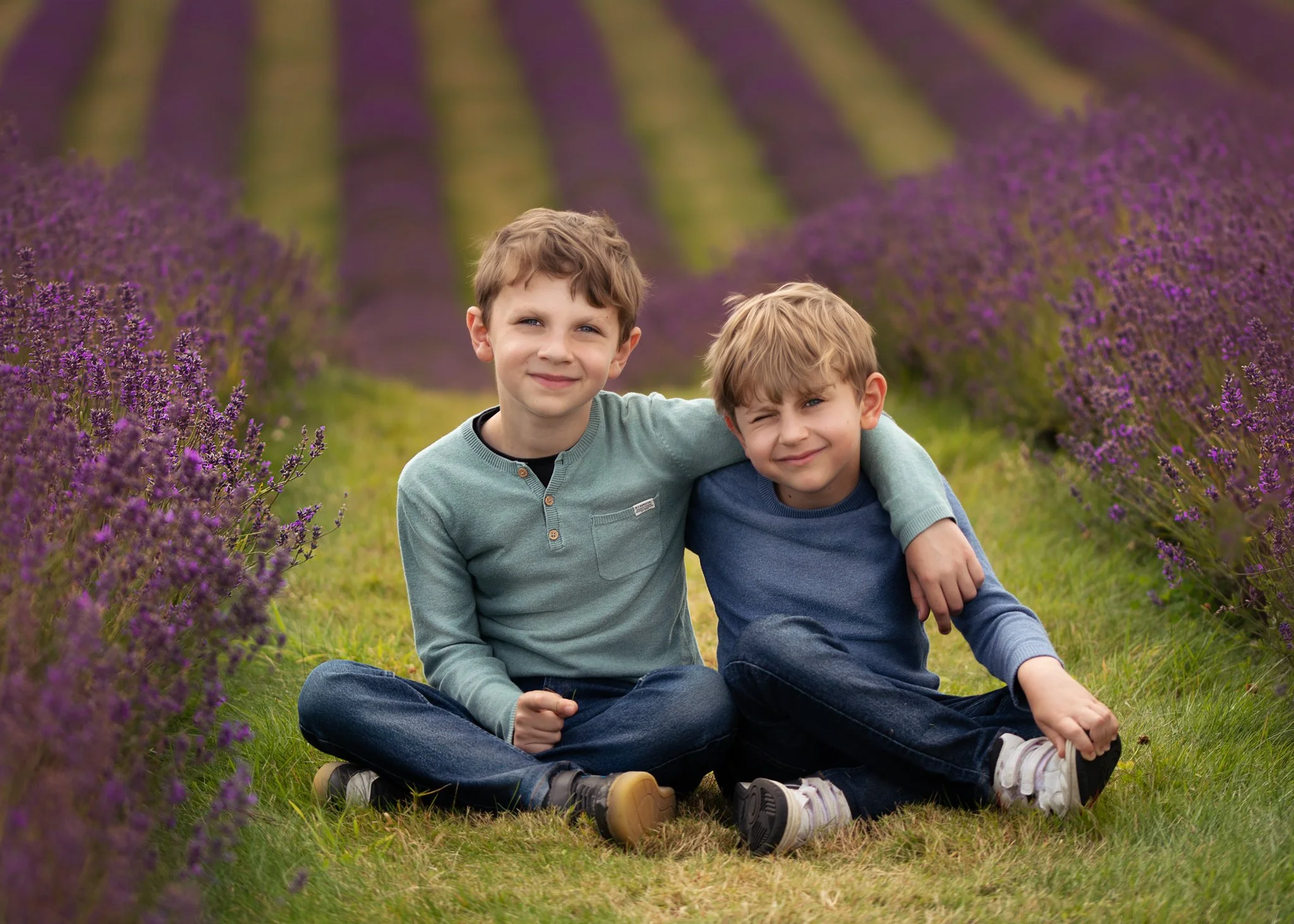 Two young boys sitting on grass among purple flowering plants, smiling with arms around each other.