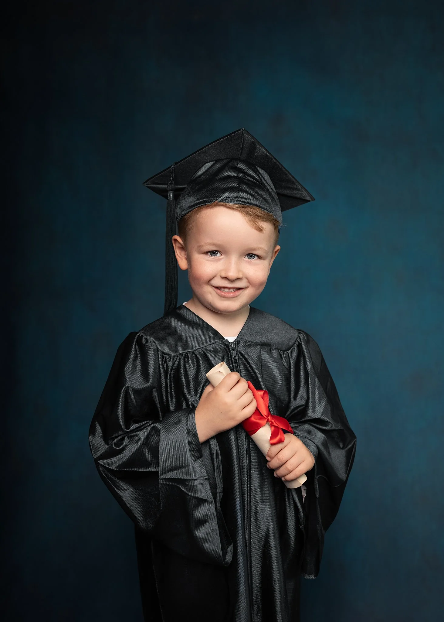 Young boy in graduation cap and gown holding a rolled diploma with a red ribbon, smiling