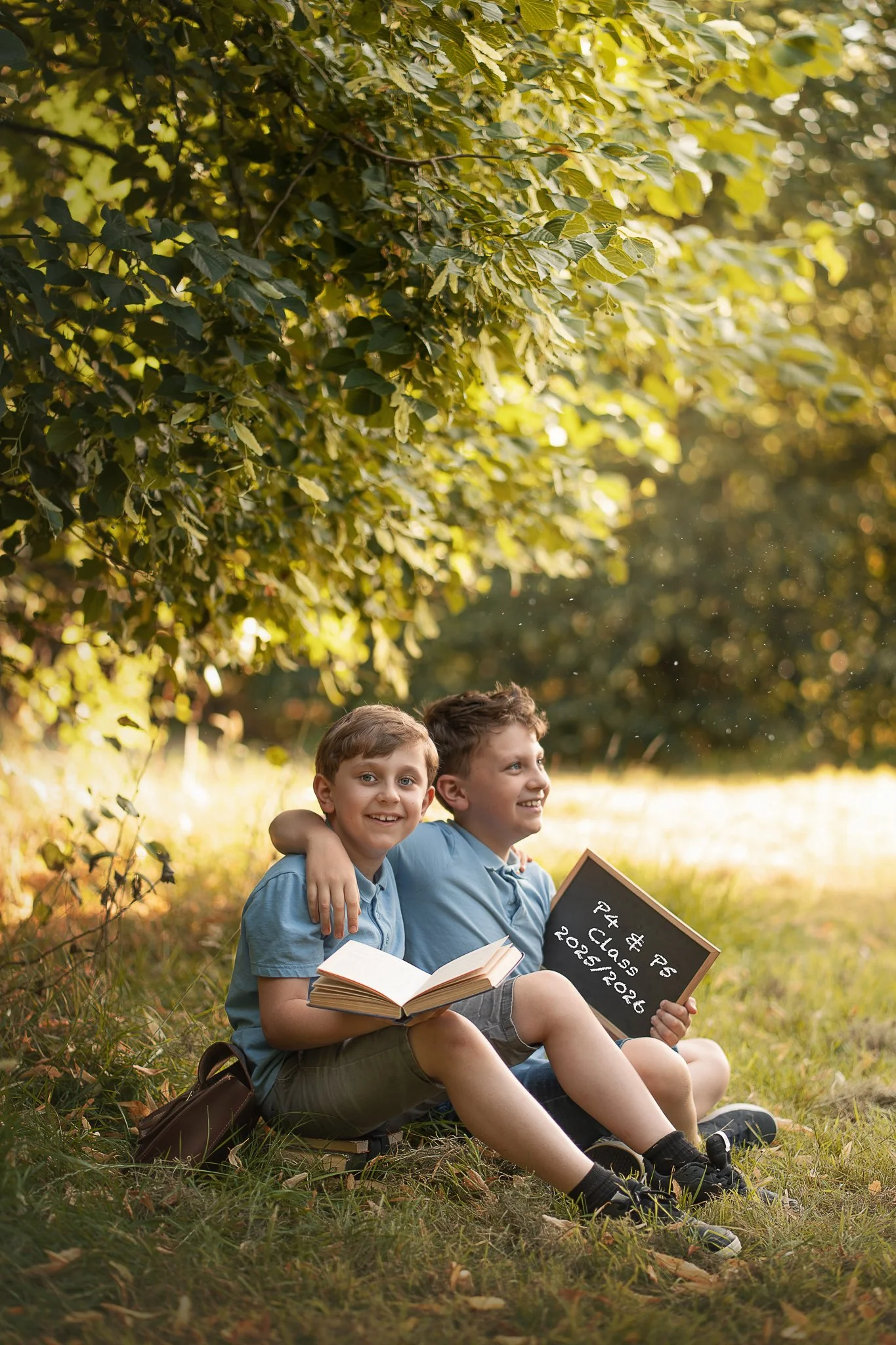 Two young boys are sitting on grass in a park, smiling and hugging each other, under a canopy of green leaves. They are holding books and a black chalkboard sign reads, '$75, 4th Grade, 2025/2026.'