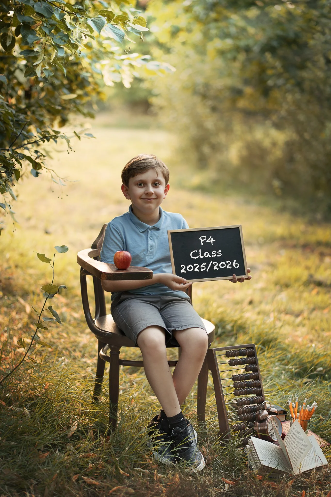 A young boy sitting outdoors in a chair surrounded by trees, holding a chalkboard with class and year information, next to books, a clock, and school supplies, with an apple on the armrest.