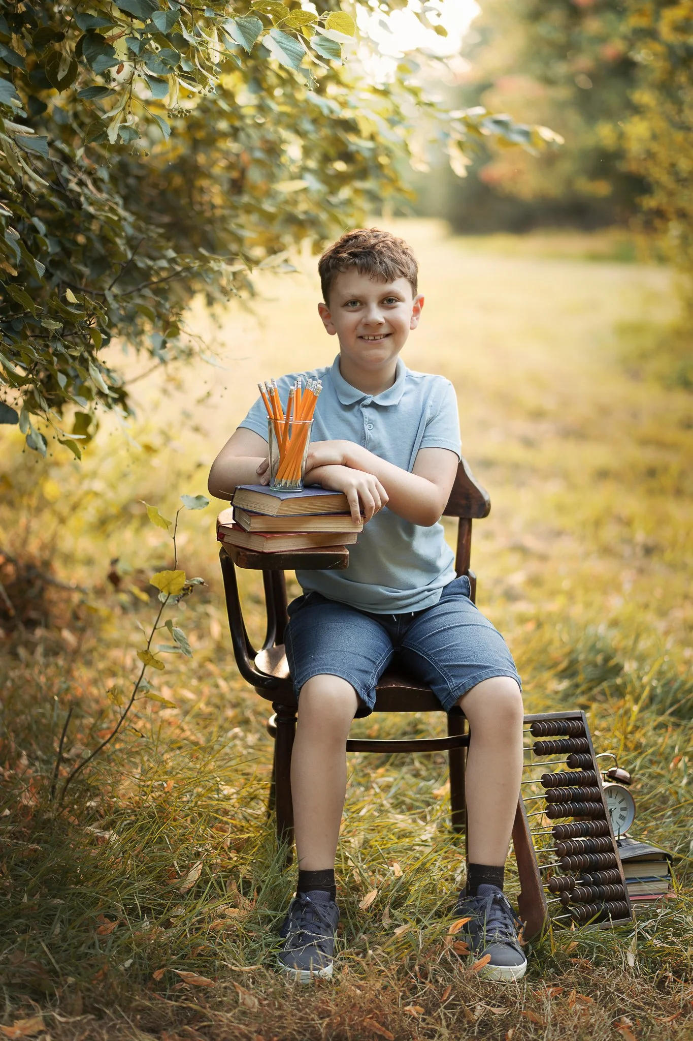 A young boy sitting outdoors on a wooden chair in a park during fall, smiling while holding a stack of books with a glass of pencils on top. There is an abacus and a clock on the ground beside him, surrounded by yellow and green leaves.