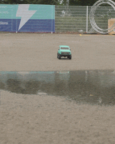 Small mint green toy car on a gravel surface near a body of water with a chain-link fence and large circular metal structure in the background.