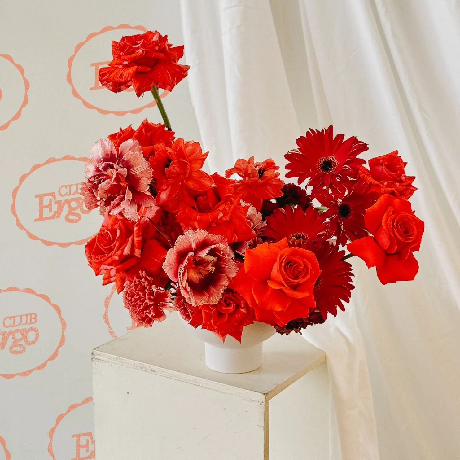 A bold floral arrangement of red and pink flowers, including roses and gerbera daisies, sits in a white vase on a pedestal, with a "Club Ergo" logo backdrop and draped fabric.