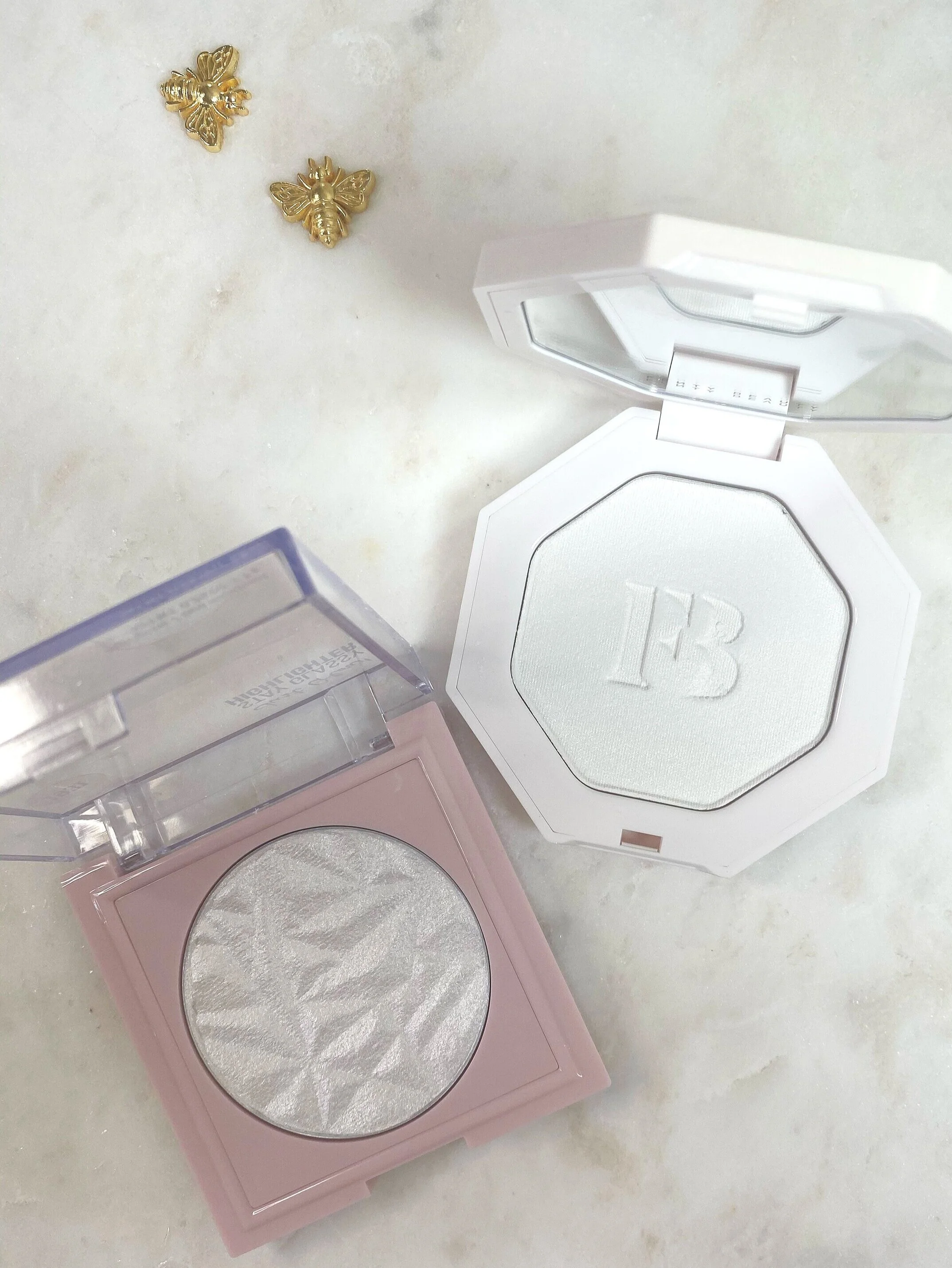 Three makeup highlighters with different shades of powder and two decorative gold bee and butterfly pins on a light surface.