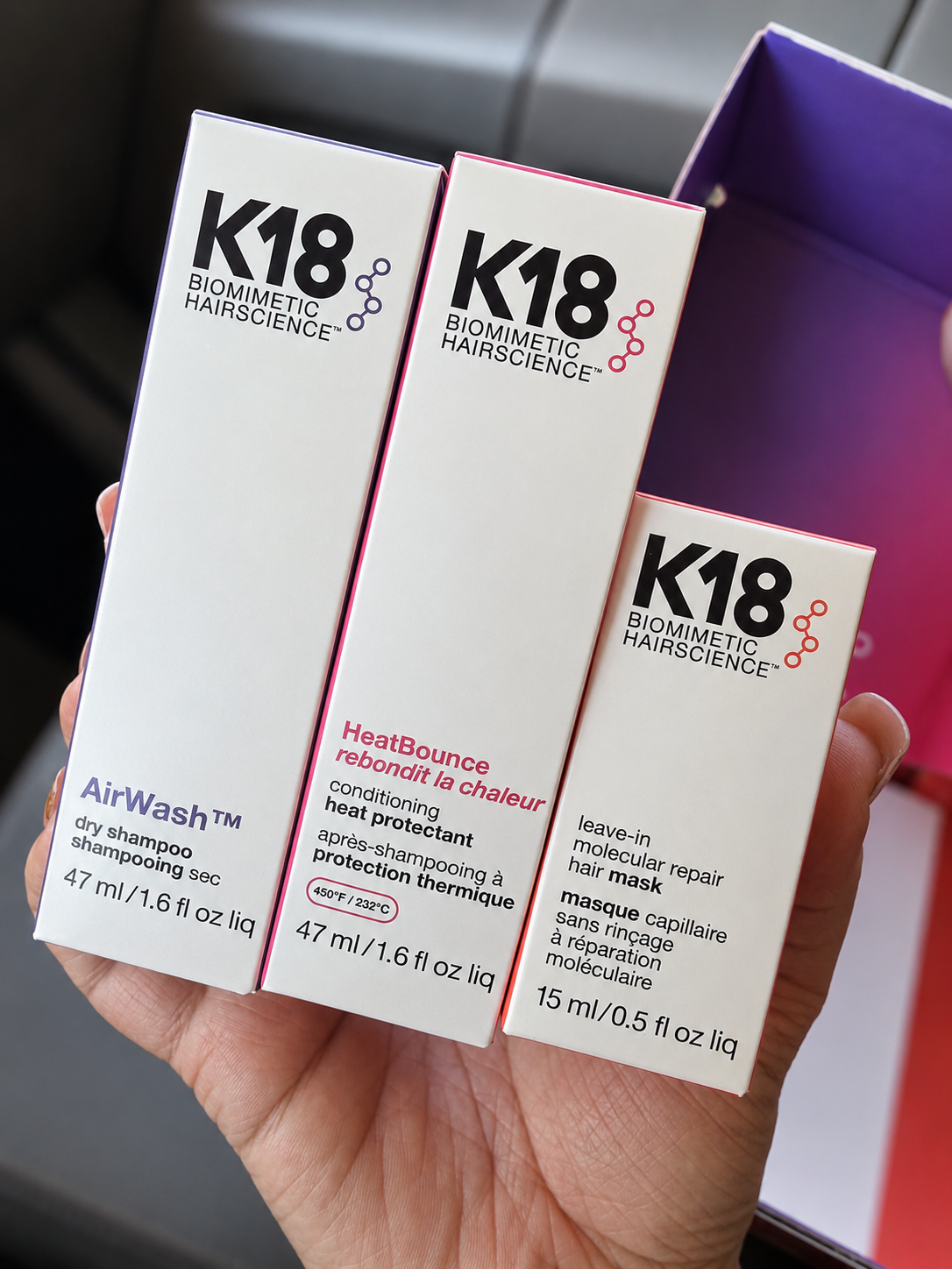 Hand holding three hair care product boxes labeled K18, including an AirWash dry shampoo, HeatBounce conditioning heat protectant, and a leave-in molecular repair hair mask.