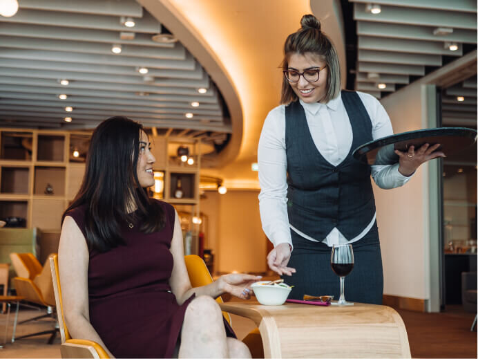 Photography capturing a smiling waitress delivering food and drink to a customer inside Manchester Airport’s premium 1903 Lounge