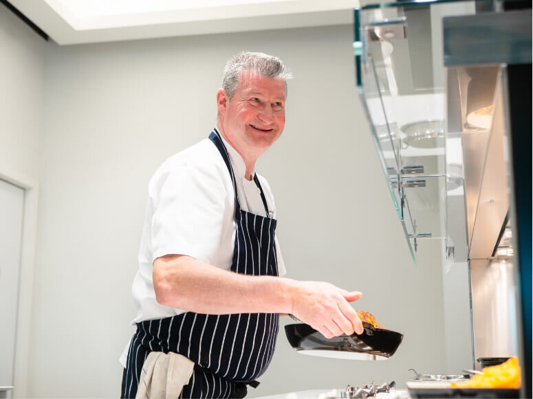 Smiling chef holding a dish of food while restocking a breakfast buffet at Manchester Airports Escape Lounges