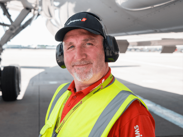 Smiling runway worker in a high vis jacket, standing under an airplane