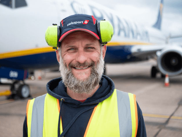 Staff portrait photography showing a smiling man and wearing noise cancelling headphones, standing next to an airplane