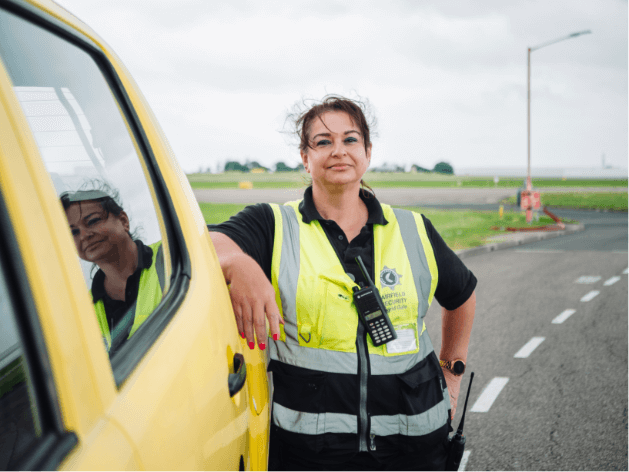 Empowered woman wearing a high vis jacket and leaning against a car next to an airport runway