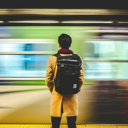 Rear view of a man in a tan coat at a subway station, with a fast-moving train blurred in motion