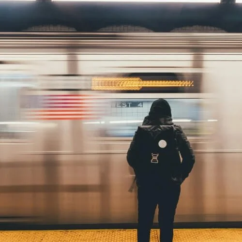 Person with a backpack standing at a subway platform, with a high-speed blurred train displaying the American flag
