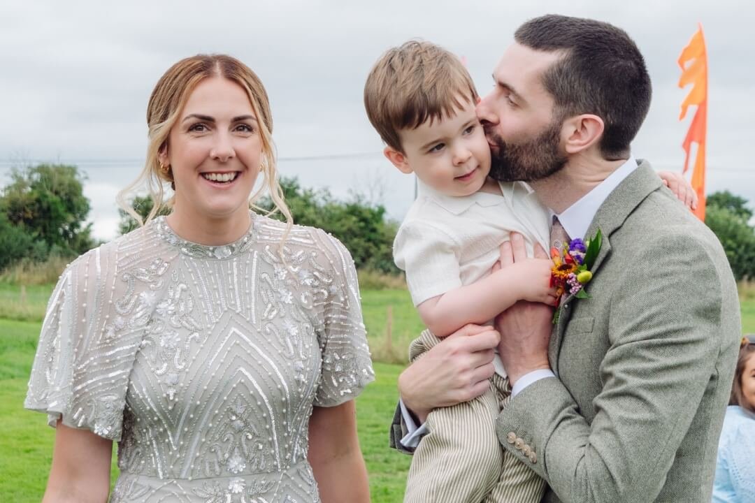 Bride in beaded dress and groom holding their son and kissing him, outdoors at wedding