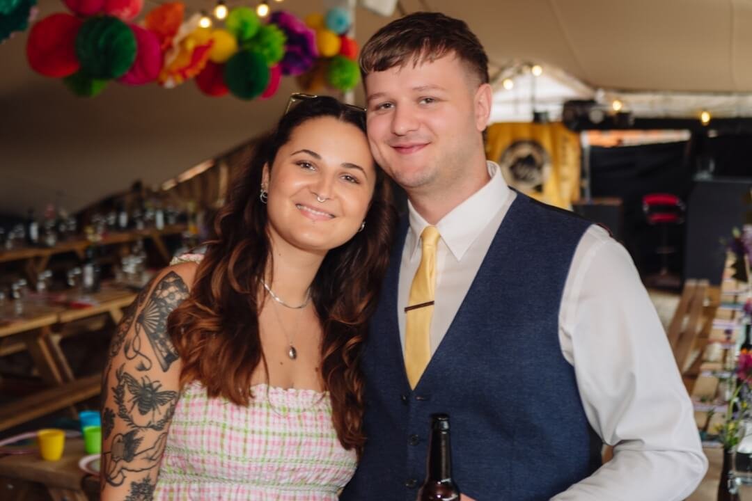 Tattooed woman and man in vest smiling and holding drinks at wedding