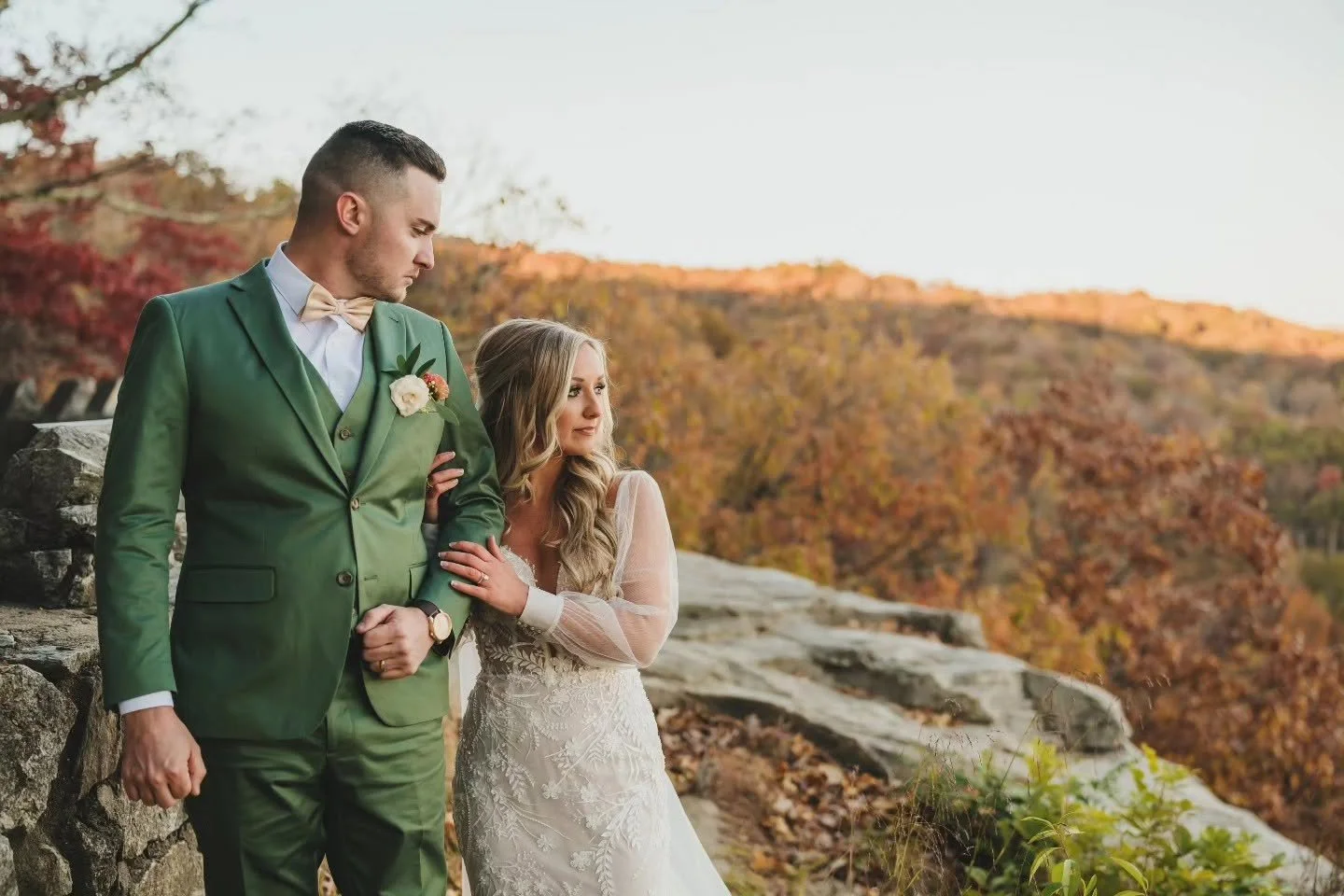 Bride in a lace wedding gown holding the arm of a groom in a green suit beside a stone wall at Pretty Place, with autumn foliage behind them.