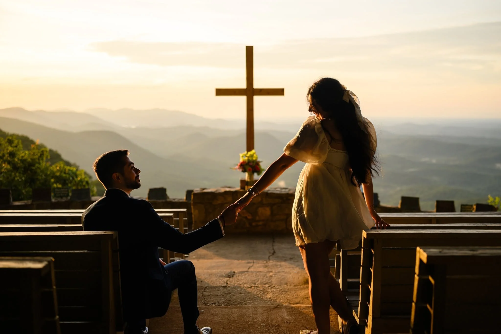 Couple holding hands between the wooden pews at Pretty Place Chapel, with the man seated and the woman standing against a sunlit mountain backdrop.