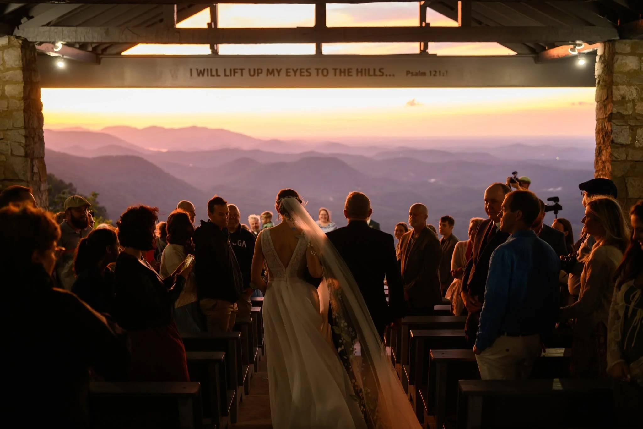 Bride walking down the aisle with her escort at Pretty Place Chapel, with guests standing on both sides and glowing skies over the mountains ahead.