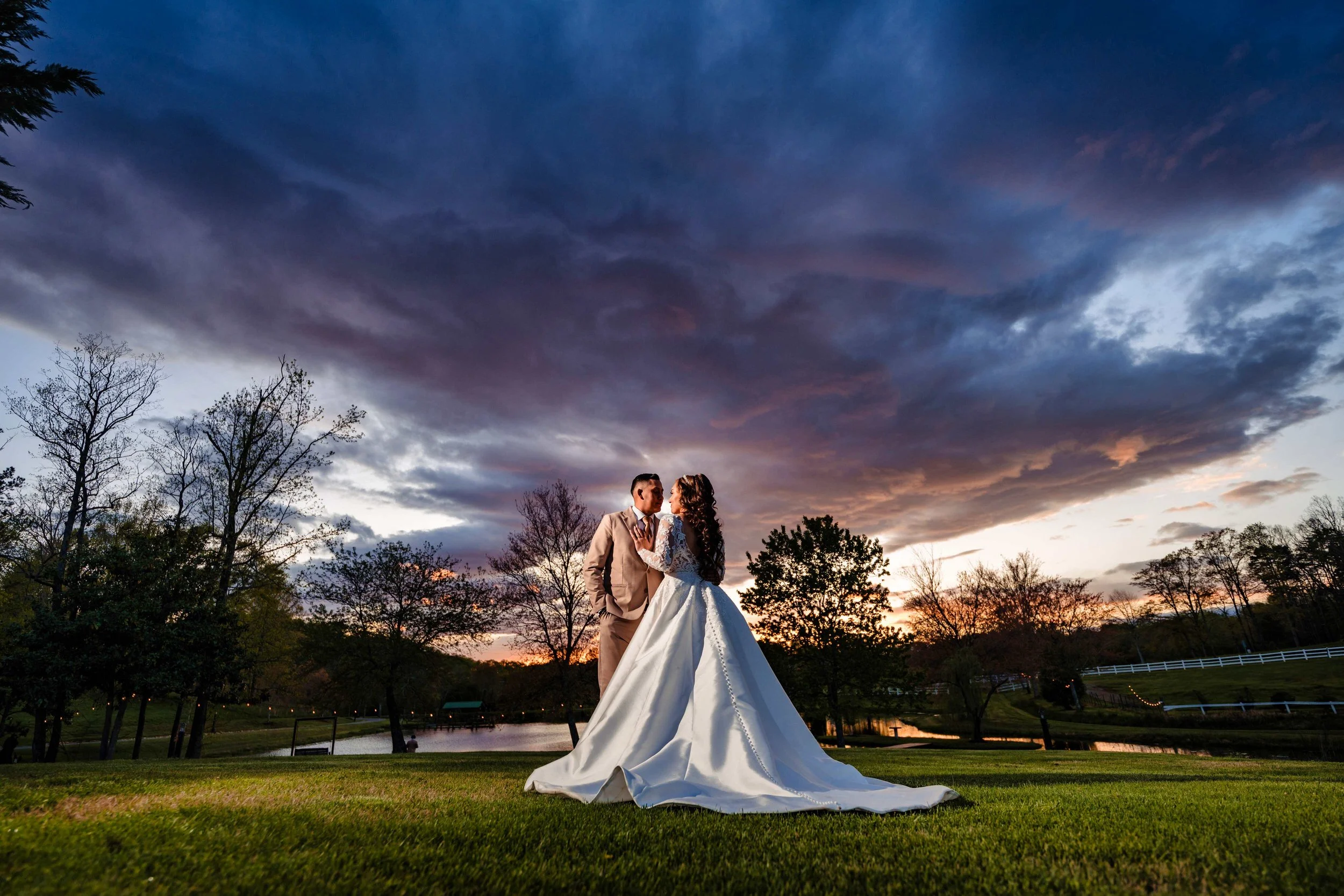 Bride and groom facing each other on the lawn at South Wind Ranch beneath sweeping blue and pink sunset clouds, with the pond and trees behind them.
