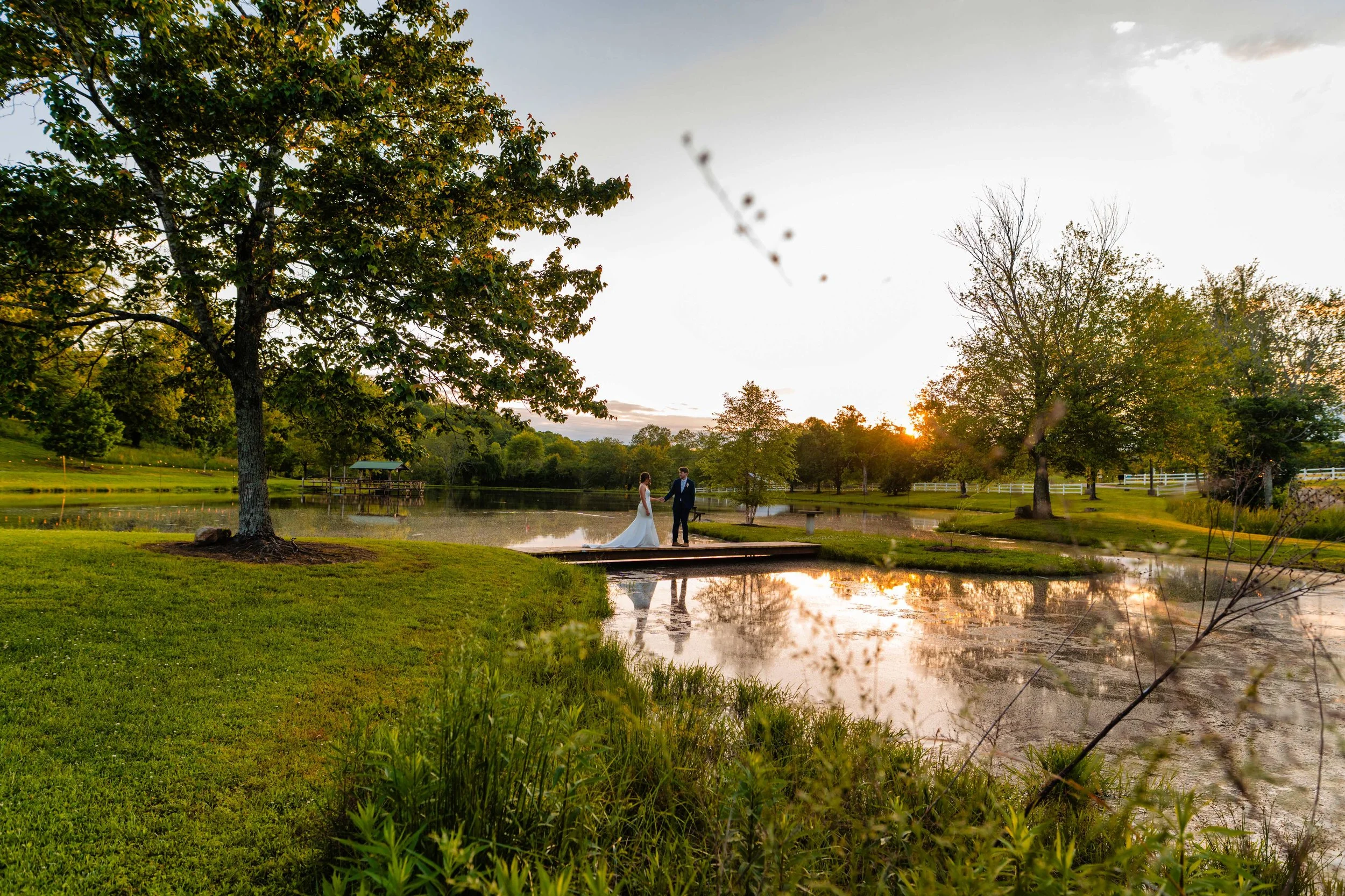 Wide view of a bride and groom holding hands on a wooden bridge at South Wind Ranch, with sunset light reflected in the pond and trees surrounding the water.
