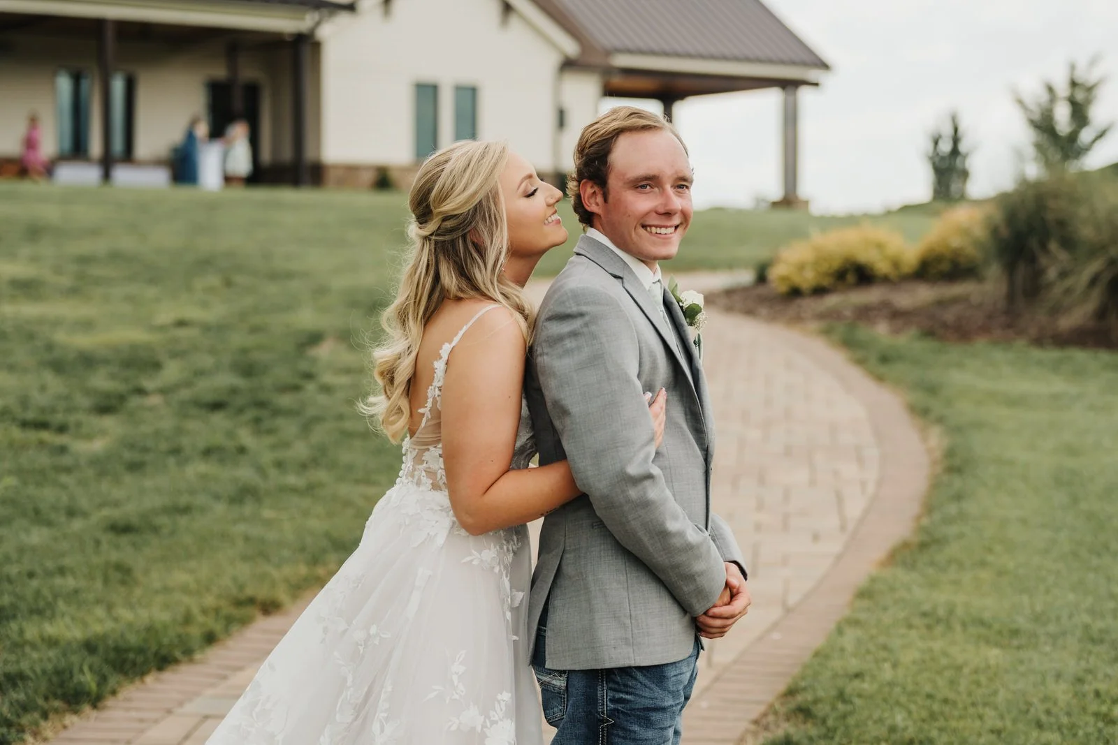 Bride hugging a smiling groom from behind on MeadowView’s curved paved pathway, with the reception building and lawn in the background.