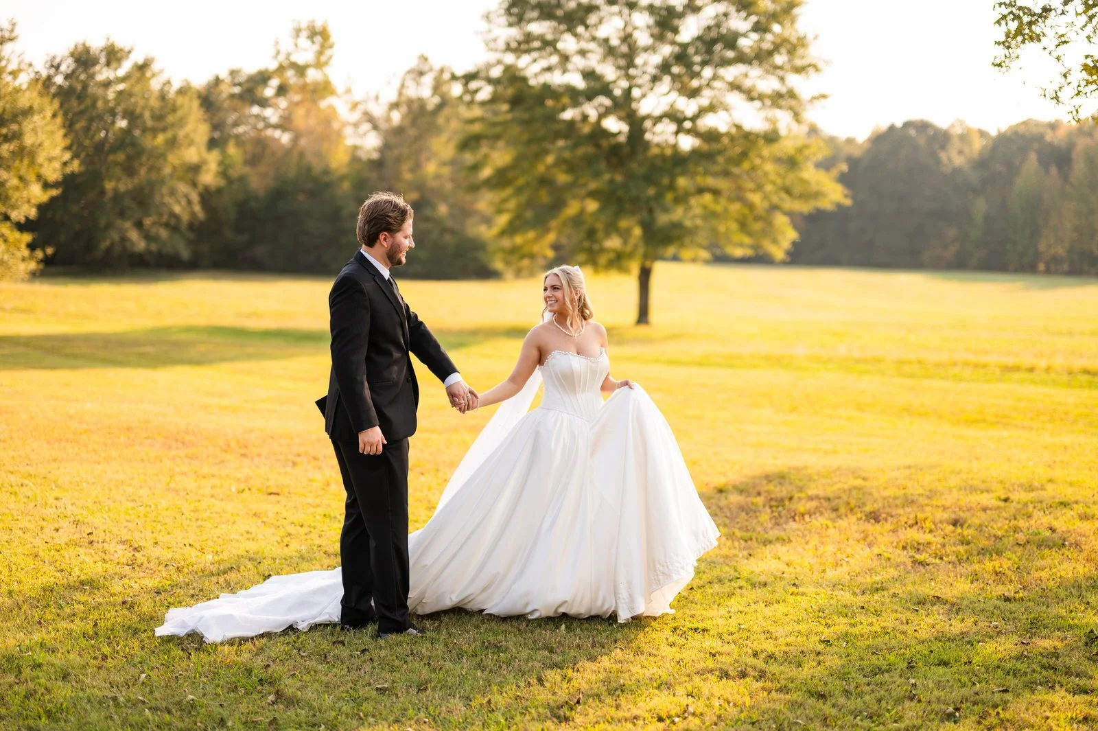 Bride and groom holding hands and smiling at each other in a sunlit field at The Barn at Sitton Hill, with her white train spread across the grass.