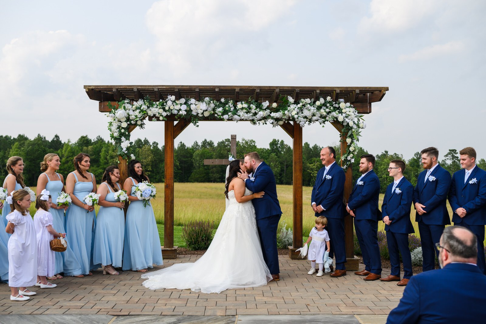 Bride and groom sharing their first kiss beneath MeadowView’s wooden pergola decorated with white flowers, surrounded by bridesmaids in light blue and groomsmen in navy suits.