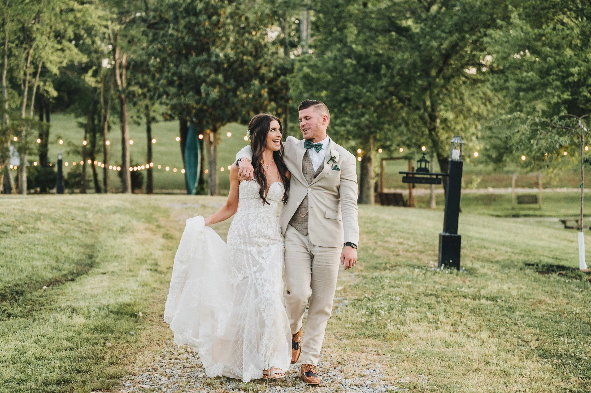 Bride and groom laughing together while walking along a gravel path at South Wind Ranch, with the groom’s arm around the bride and string lights glowing behind them.