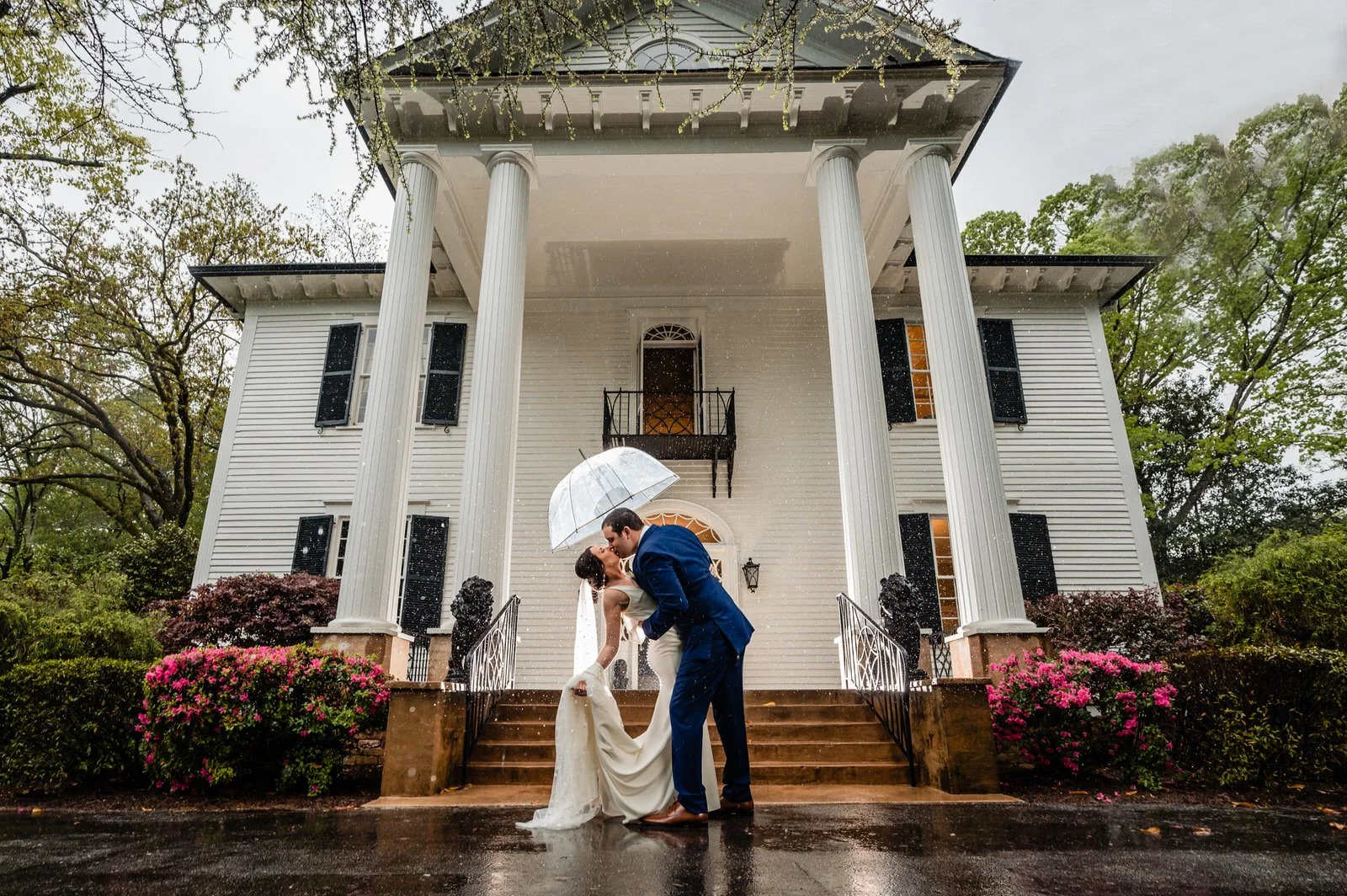 Bride and groom kissing beneath a clear umbrella in the rain outside Duncan Estate’s white-columned house in Spartanburg, SC.