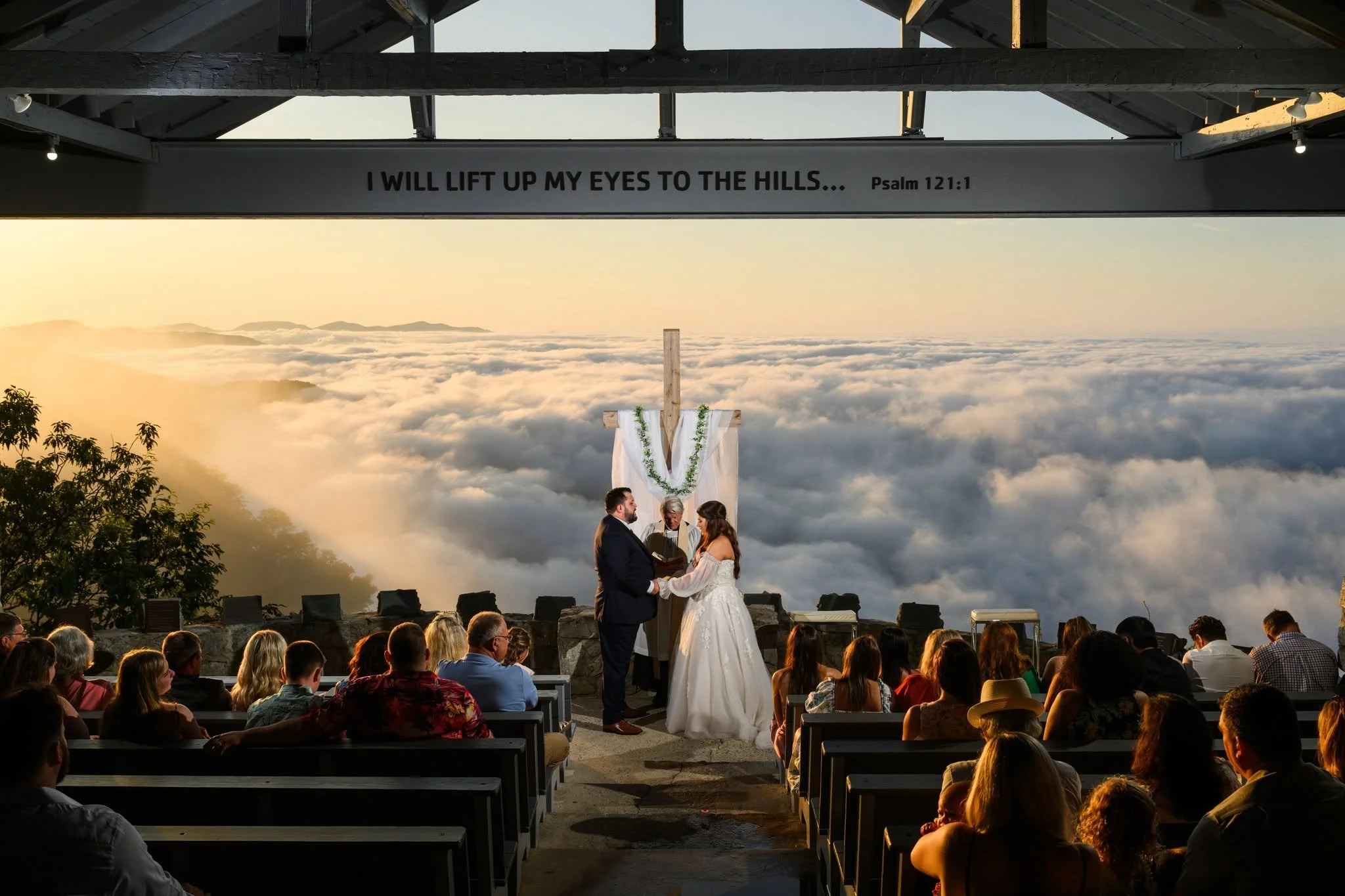 Bride and groom holding hands during their sunrise wedding ceremony at Pretty Place Chapel, with clouds filling the mountain valley behind them.