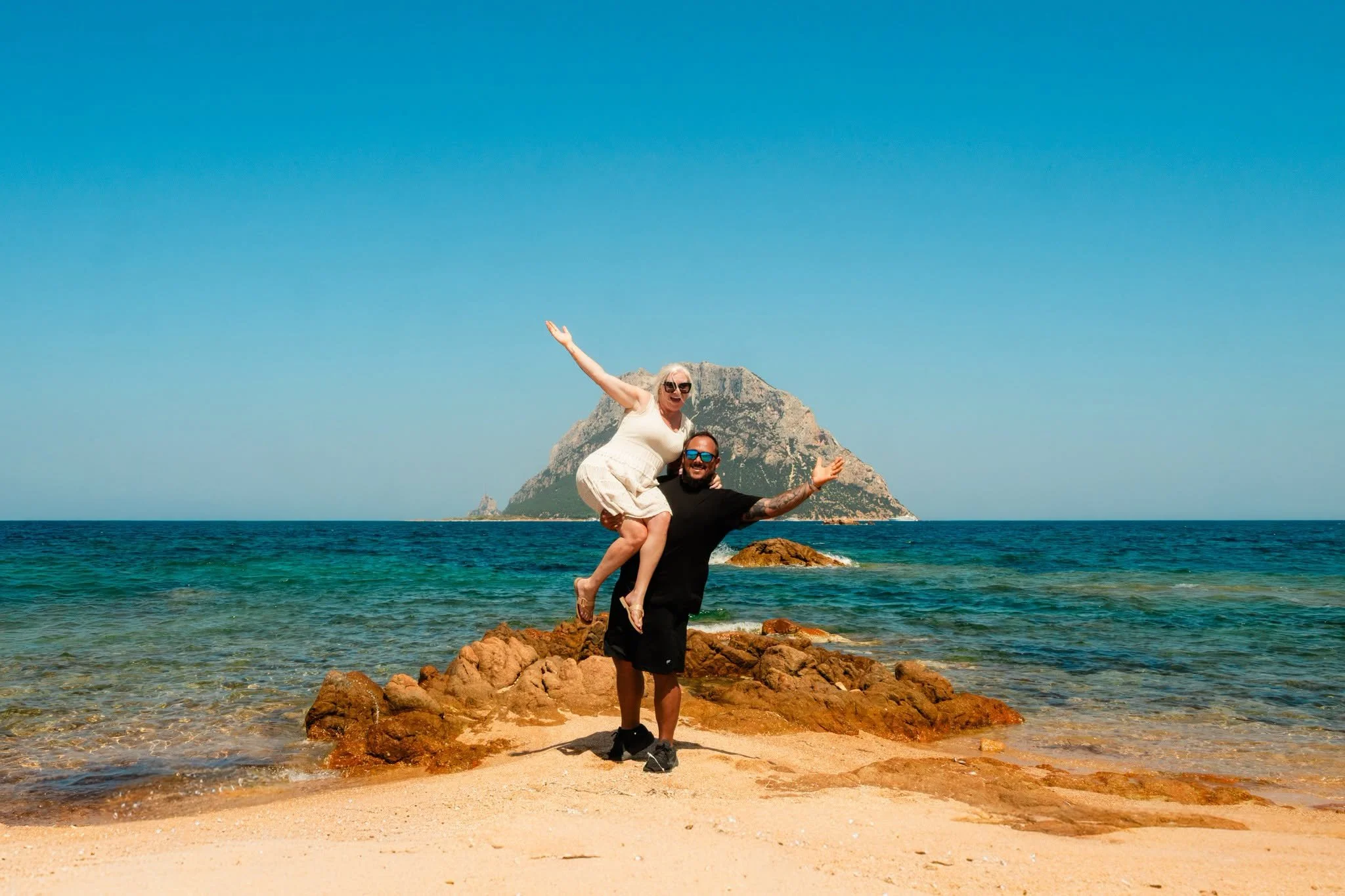 Brandon lifting Katie on a rocky beach, with turquoise water and a mountainous island behind them.