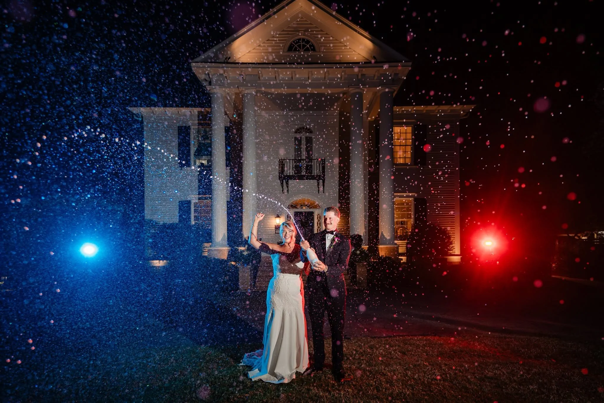 Bride and groom celebrating with a champagne spray outside Duncan Estate at night, with red and blue lighting illuminating the droplets and historic house.