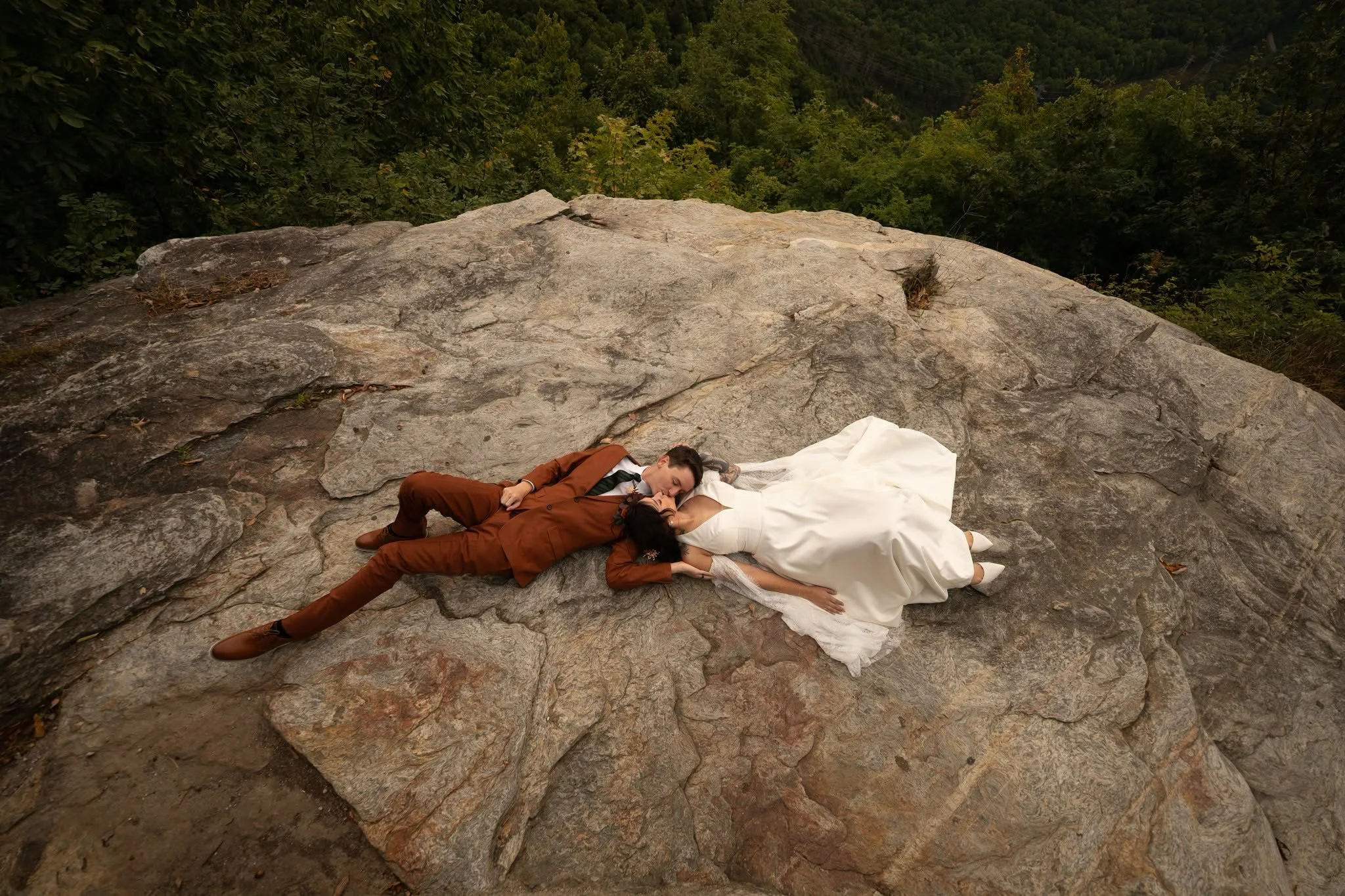 Bride in a white gown and groom in a rust-colored suit lying together and kissing on a broad rock surrounded by forest near Pretty Place Chapel.