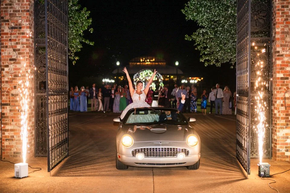 Bride raising her arms as she and the groom leave Edinburgh West in a silver convertible, passing between ornate iron gates and spark fountains while guests watch behind them.