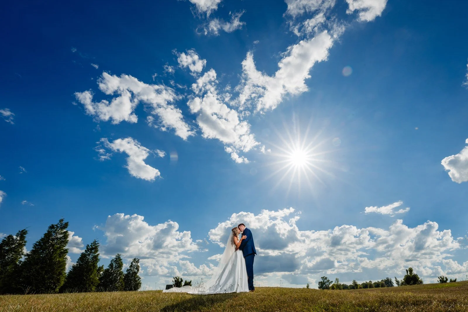 Bride and groom kissing on MeadowView’s grassy hillside beneath a vivid blue sky, white clouds, and a bright sunburst.