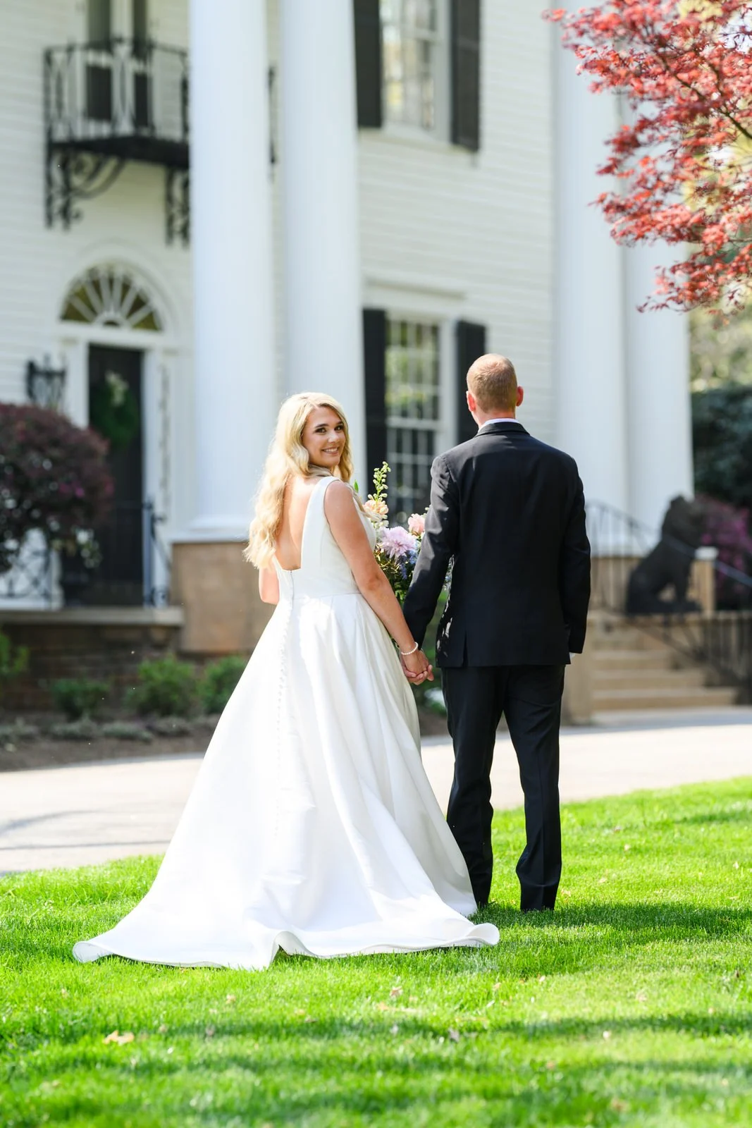 Bride looking back at the camera while holding the groom’s hand on Duncan Estate’s front lawn in Spartanburg, SC, with the historic house behind them.