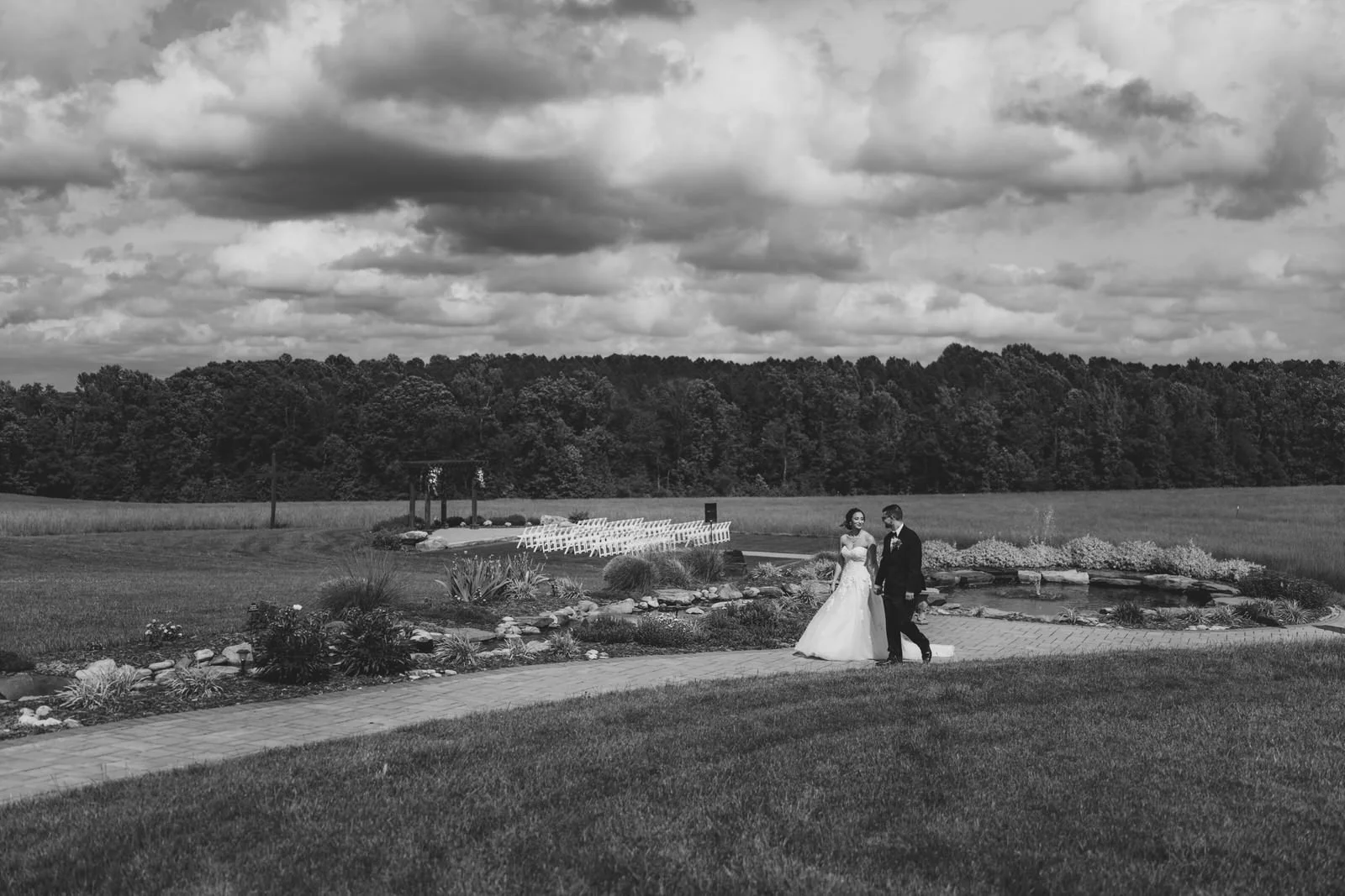 Black and white photo of a bride and groom walking hand in hand along MeadowView’s garden path, with a pond, outdoor ceremony seating, and dramatic clouds behind them.