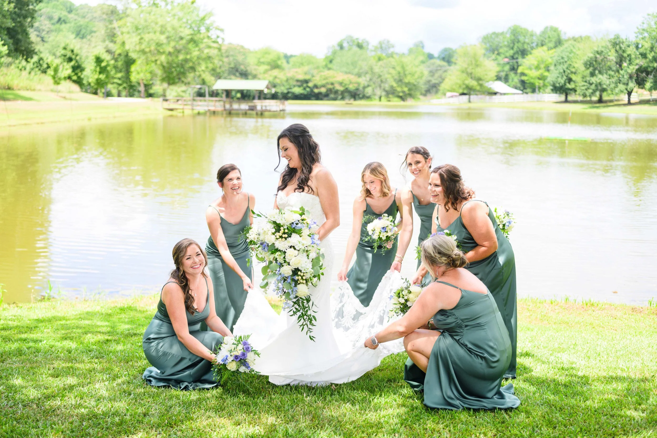 Bride laughing with her bridesmaids in green dresses as they arrange her wedding gown beside the pond at South Wind Ranch.